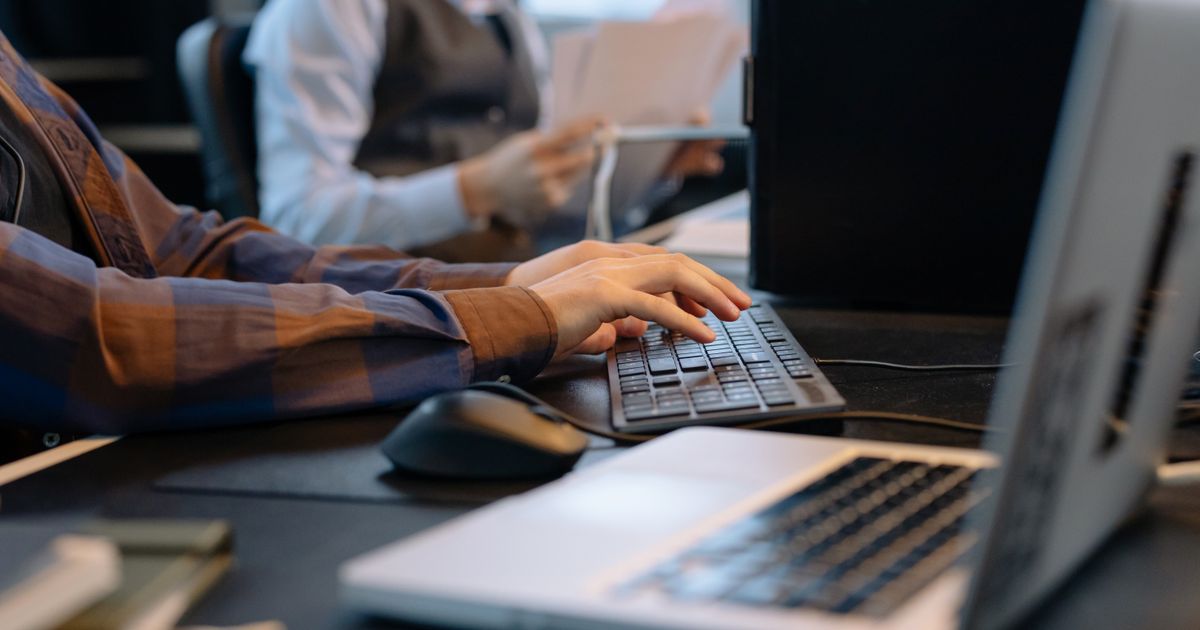 A person typing at a computer keyboard while a colleague sorts paperwork nearby